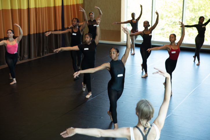 Group of dancers in the same pose during a dance workshop in a brightly lit studio space. They are all standing with one arm out to the side and another gracefully above their heads